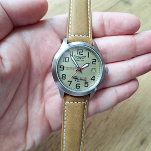 K&ouml;nnte beinhalten: Eine braune Lederarmbanduhr mit einem silbernen Geh&auml;use und einem wei&szlig;en Zifferblatt. Das Zifferblatt hat schwarze Zahlen und Zeiger, einen roten Sekundenzeiger und eine kleine Flugzeuggrafik. Das Zifferblatt der Uhr hat auch den Text "Авиатор" und "Гастелло Н. Ф. 1941 год СССР".