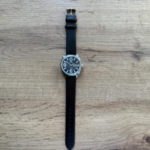 May include: A black wristwatch with a silver-colored case and a black leather band. The watch face is black with white numerals and hands. The watch is displayed on a wooden surface.