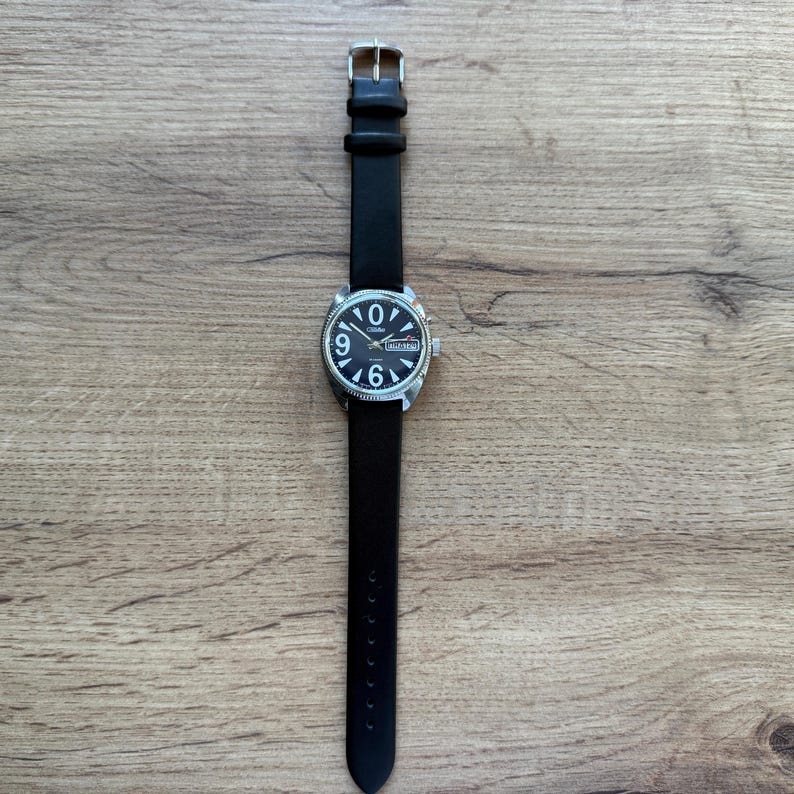 May include: A black wristwatch with a silver-colored case and a black leather band. The watch face is black with white numerals and hands. The watch is displayed on a wooden surface.