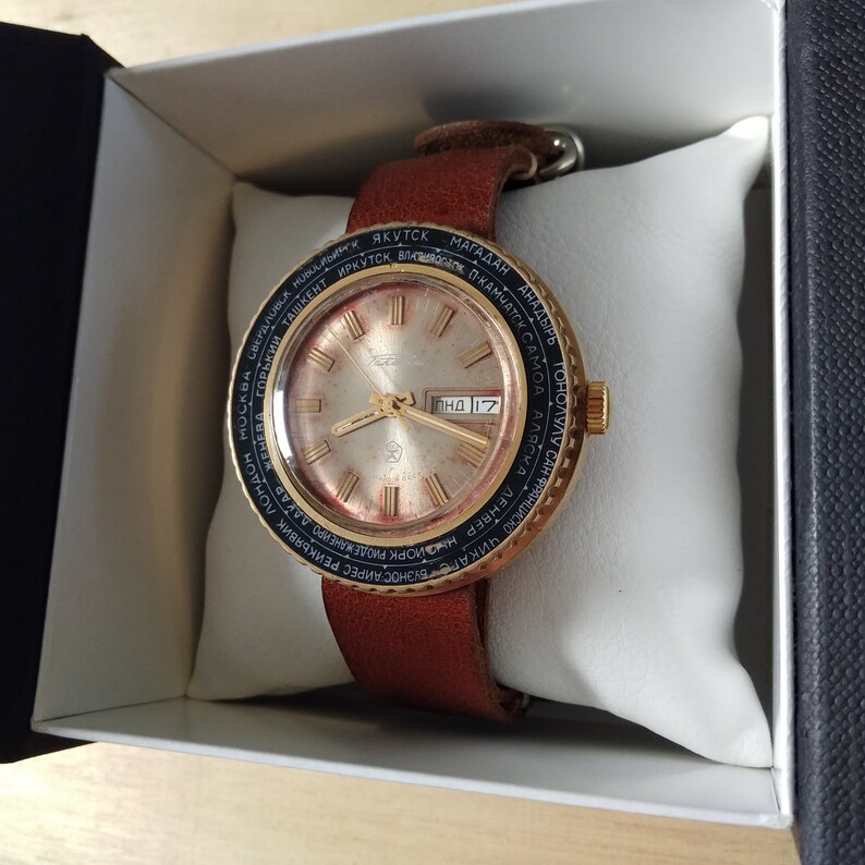 Puede incluir: Un reloj de pulsera dorado con un bisel negro y dorado. La esfera del reloj es blanca con marcas negras y una ventana de fecha que indica "ПНД 17". El reloj est&aacute; en una correa de cuero marr&oacute;n.