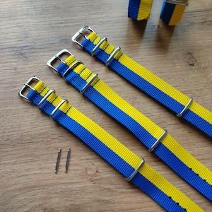 May include: Three yellow and blue striped watch straps with silver buckles. The straps are laid out on a wooden surface.