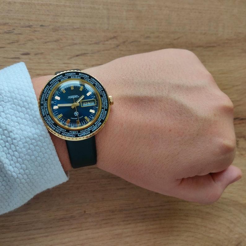 May include: A gold-tone wristwatch with a black face and a black leather band. The watch face has a world time dial with city names in Cyrillic and Latin script. The watch face also has a date window and a second hand.