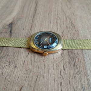 Puede incluir: Un reloj de pulsera Sekonda vintage con caja dorada y correa de malla. La esfera del reloj es marr&oacute;n oscuro con agujas e indicadores dorados, y una ventana de fecha a las 6. El reloj est&aacute; sobre un fondo de madera.