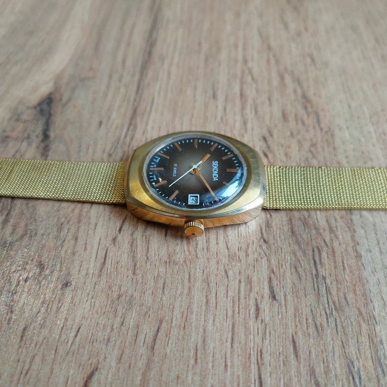 Puede incluir: Un reloj de pulsera Sekonda vintage con caja dorada y correa de malla. La esfera del reloj es marr&oacute;n oscuro con agujas e indicadores dorados, y una ventana de fecha a las 6. El reloj est&aacute; sobre un fondo de madera.