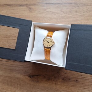 Op de afbeelding: Een goudkleurig polshorloge met een lichtbruine leren band, gepresenteerd in een zwart-witte geschenkdoos. De wijzerplaat van het horloge heeft gouden wijzers en uurmarkeringen. Het horloge ligt op een wit kussen.