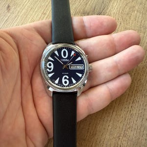 May include: A silver-toned wristwatch with a black leather band. The watch face is dark blue with large white numerals at 9, 6, and 0. The watch also displays the day and date. The watch is being held.