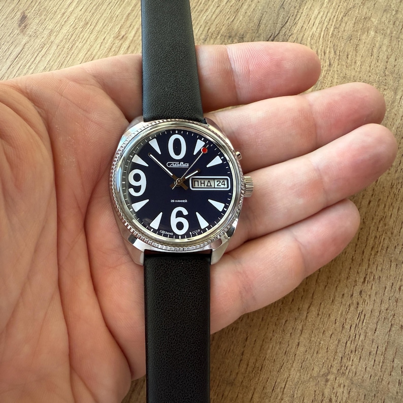May include: A silver-toned wristwatch with a black leather band. The watch face is dark blue with large white numerals at 9, 6, and 0. The watch also displays the day and date. The watch is being held.