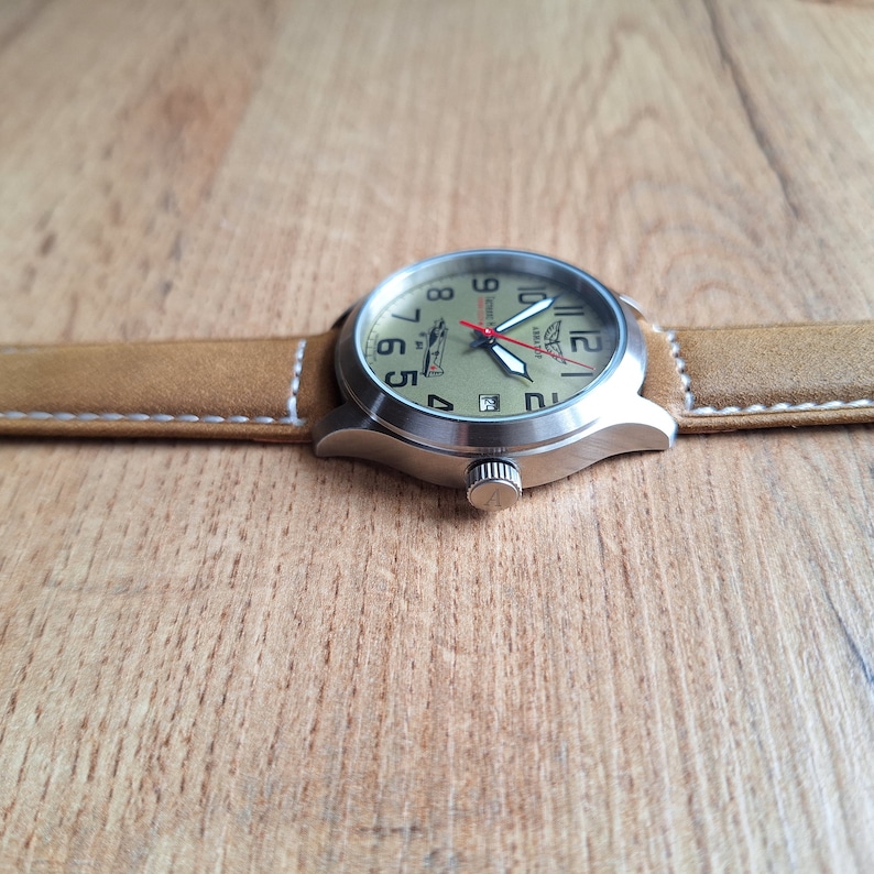 K&ouml;nnte beinhalten: Eine braune Lederarmbanduhr mit einem silbernen Geh&auml;use und einem wei&szlig;en Zifferblatt. Das Zifferblatt hat schwarze Zahlen und Zeiger sowie einen kleinen roten Sekundenzeiger. Das Zifferblatt hat auch eine kleine Flugzeuggrafik und den Text "Soviet Air Force" aufgedruckt.