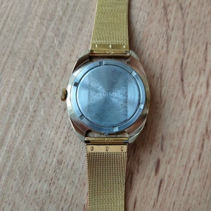 Puede incluir: Un reloj de pulsera dorado con una correa de malla. La caja del reloj es dorada y tiene una parte trasera redonda de color plateado con el n&uacute;mero "545598" impreso. La correa del reloj es una fina malla de metal dorado.