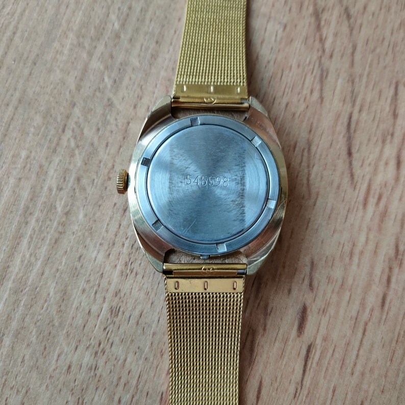 Puede incluir: Un reloj de pulsera dorado con una correa de malla. La caja del reloj es dorada y tiene una parte trasera redonda de color plateado con el n&uacute;mero "545598" impreso. La correa del reloj es una fina malla de metal dorado.