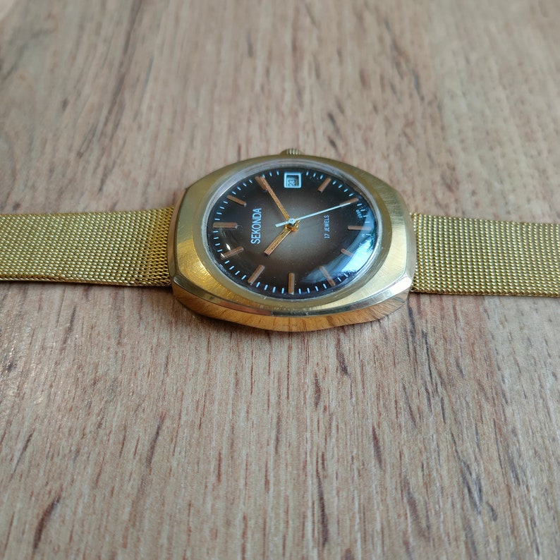 Puede incluir: Un reloj Sekonda dorado con esfera marr&oacute;n y correa de malla. La caja rectangular muestra la fecha. La esfera tiene agujas y marcadores dorados. La correa mide unos 2 cm de ancho. La esfera indica '17 Jewels'.