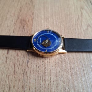 Op de afbeelding: Een gouden polshorloge met een blauwe wijzerplaat en een zwart leren bandje. De wijzerplaat van het horloge heeft een maanfase-indicator en het woord "Raketa" staat in goud gedrukt.