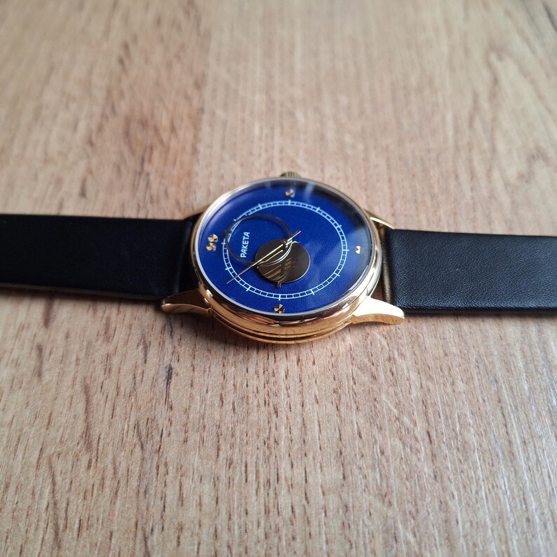 Op de afbeelding: Een gouden polshorloge met een blauwe wijzerplaat en een zwart leren bandje. De wijzerplaat van het horloge heeft een maanfase-indicator en het woord "Raketa" staat in goud gedrukt.