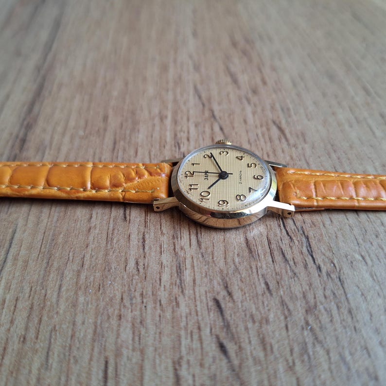 Op de afbeelding: Een vintage polshorloge met een goudkleurige kast en een lichtgekleurde wijzerplaat. Het horloge heeft zwarte cijfers en wijzers en een bruinleren band. Het horloge wordt getoond op een houten oppervlak.