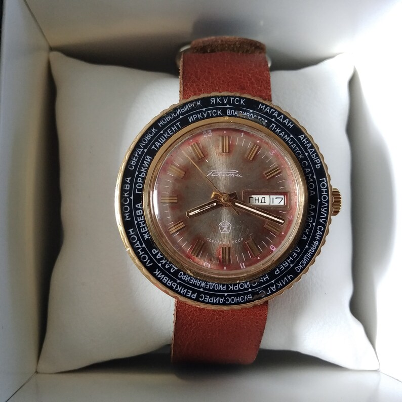 Puede incluir: Un reloj de pulsera dorado con una correa de cuero negro y una esfera redonda. La esfera tiene un fondo blanco con marcas negras y una aguja de segundos roja. El reloj tiene un bisel de zona horaria de 24 horas con nombres de ciudades impresos en negro sobre un fondo dorado. La esfera del reloj tambi&eacute;n incluye el texto "&Pi;&Eta;&Delta; 17".