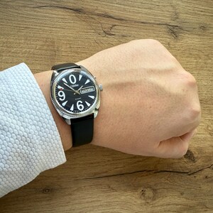 May include: A black wristwatch with a black leather band. The watch face is black with white numerals and hands. The watch has a silver-colored bezel and a date display. The watch is on a wrist.