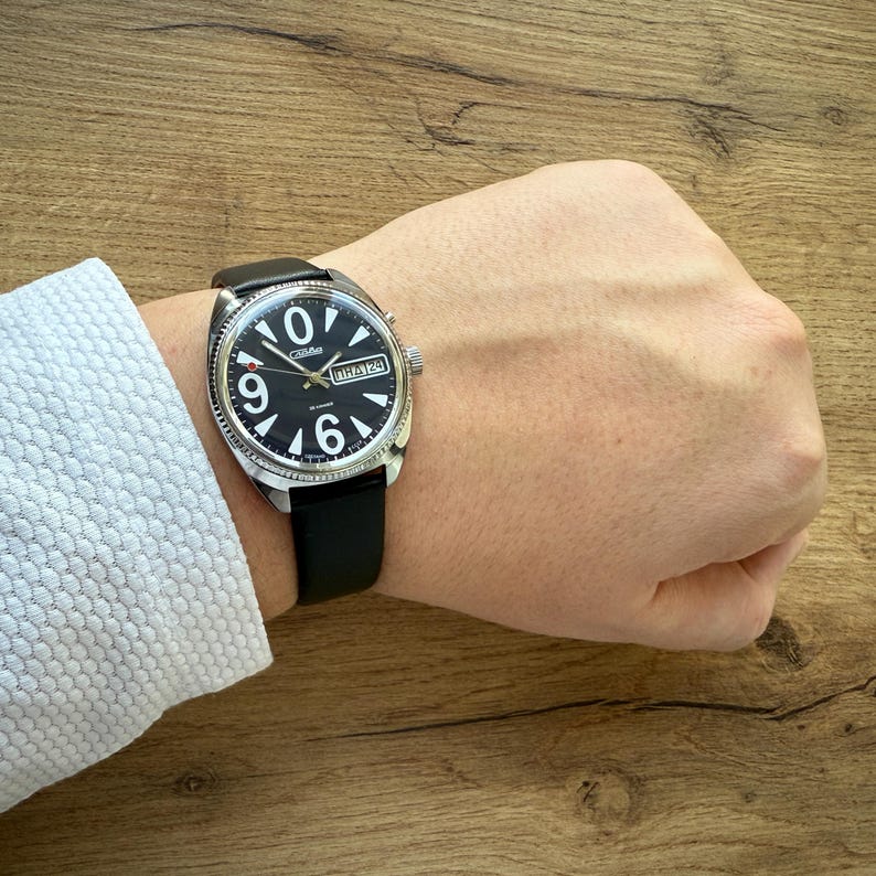 May include: A black wristwatch with a black leather band. The watch face is black with white numerals and hands. The watch has a silver-colored bezel and a date display. The watch is on a wrist.