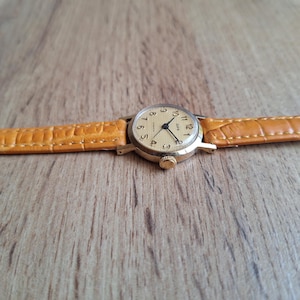 Op de afbeelding: Een vintage goudkleurig polshorloge met een lichtgekleurde wijzerplaat en zwarte wijzers. Het horloge heeft een bruin lederen bandje. De wijzerplaat toont de cijfers 1 tot en met 12. Het horloge ligt op een houten achtergrond.