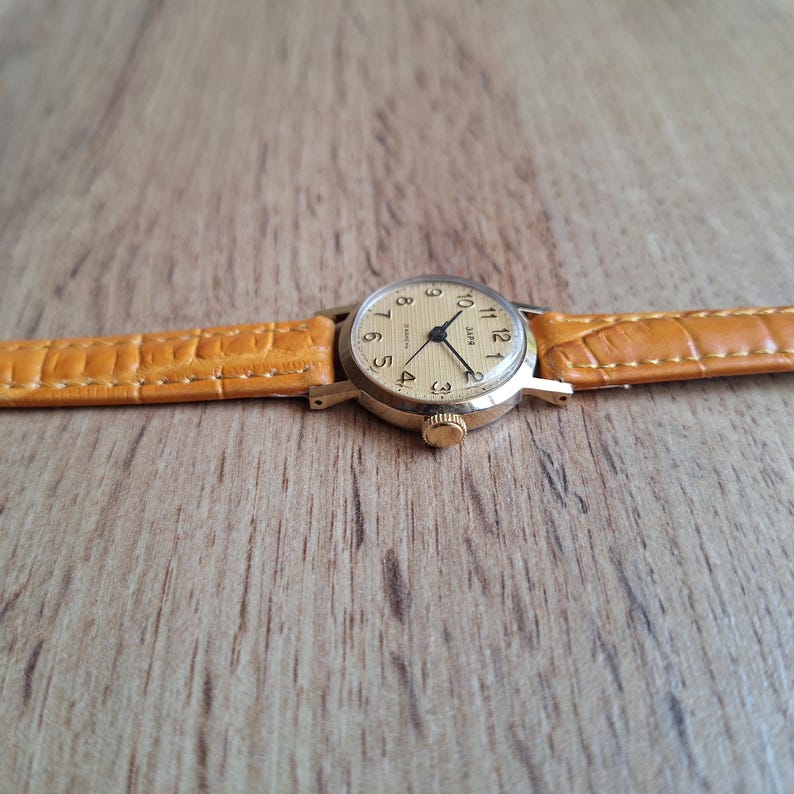Op de afbeelding: Een vintage goudkleurig polshorloge met een lichtgekleurde wijzerplaat en zwarte wijzers. Het horloge heeft een bruin lederen bandje. De wijzerplaat toont de cijfers 1 tot en met 12. Het horloge ligt op een houten achtergrond.