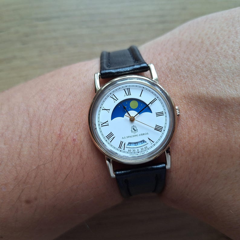 Op de afbeelding: Een goudkleurig polshorloge met een witte wijzerplaat, zwarte Romeinse cijfers en een maanfase-indicator. Het horloge heeft een zwarte leren band en de tekst "A.G. Spalding & Bros." is zichtbaar op de wijzerplaat.