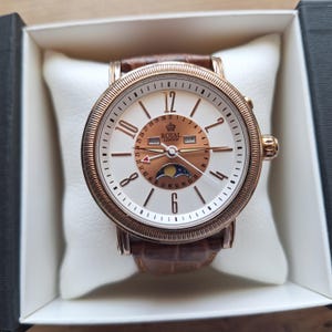 Peut inclure: Une montre Royal London dorée avec un cadran blanc, un bracelet en cuir marron et un indicateur de phase lunaire. La montre est présentée dans une boîte blanche avec un bord noir. Le cadran de la montre porte l'inscription "ROYAL LONDON".