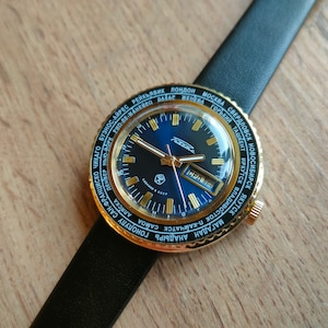 May include: A gold-toned wristwatch with a blue face and a black leather band. The watch face has a world time dial with city names in Cyrillic and Latin script. The watch face also has a date window and a day of the week window. The watch is made in the USSR.