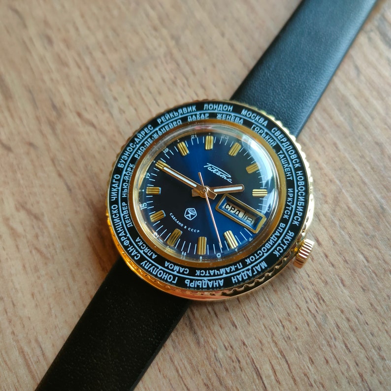 May include: A gold-toned wristwatch with a blue face and a black leather band. The watch face has a world time dial with city names in Cyrillic and Latin script. The watch face also has a date window and a day of the week window. The watch is made in the USSR.