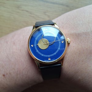 Op de afbeelding: Een gouden polshorloge met een blauwe wijzerplaat en een zwarte leren band. De wijzerplaat van het horloge heeft een gouden cirkel met een kleinere gouden cirkel erin. De tekst "PAKEITA" is zichtbaar op de wijzerplaat van het horloge.
