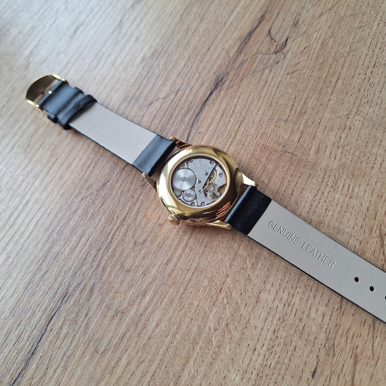 Op de afbeelding: Een gouden polshorloge met een zwart leren bandje. De wijzerplaat van het horloge is zichtbaar, waardoor het binnenwerk van het horloge te zien is. Het bandje is gemarkeerd met "GENUINE LEATHER".