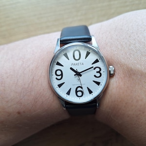 May include: A silver wristwatch with a white face and black numerals 0, 3, 6, and 9. The watch has a black leather strap and the brand name "Paketa" is displayed on the face. The watch is on a wrist.