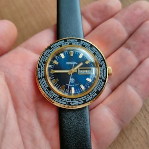 May include: A gold-toned wristwatch with a blue face and a black leather band. The watch face features a world map with city names printed around the edge. The watch face also includes a date window and a day of the week window. The watch is made by the brand "Poljot".