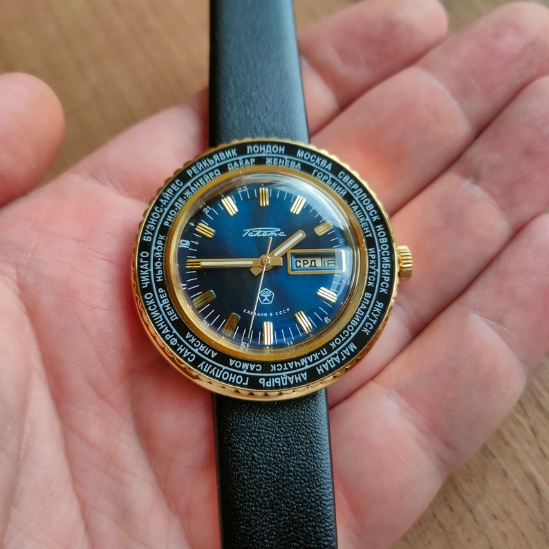 May include: A gold-toned wristwatch with a blue face and a black leather band. The watch face features a world map with city names printed around the edge. The watch face also includes a date window and a day of the week window. The watch is made by the brand "Poljot".