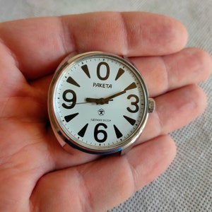 May include: A circular watch face with a silver casing and a white dial. Large black numerals indicate the hours, with the brand name "RAKETA" at the top. The watch has black hands and the text "СДЕЛАНО В СССР" at the bottom.
