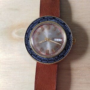 Puede incluir: Un reloj de pulsera dorado con una esfera negra y una correa de cuero marr&oacute;n. La esfera del reloj tiene un mapa del mundo impreso alrededor del borde con nombres de ciudades en escritura cir&iacute;lica. La esfera del reloj tambi&eacute;n tiene una ventana de fecha y una ventana de d&iacute;a de la semana. El reloj est&aacute; configurado en "ПНД 17".