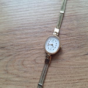 May include: A vintage gold-toned wristwatch with an oval white face. The watch features the text "ЧАЙКА" and "17 камней" on the face. The watch has a braided gold band. The watch is displayed on a wooden surface.