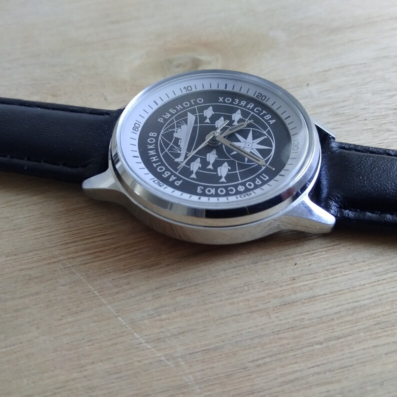 Puede incluir: Un reloj de pulsera plateado con una esfera negra y una correa de cuero negra. La esfera del reloj presenta una br&uacute;jula, un barco y peces. El texto en la esfera dice "ПРОФСОЮЗ3 РАБОТНИКОВ РЫБНОГО ХОЗЯЙСТВА 1991".
