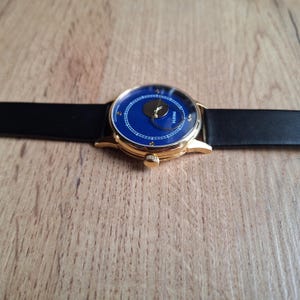 Op de afbeelding: Een gouden polshorloge met een blauwe wijzerplaat en een zwarte leren band. De wijzerplaat van het horloge heeft een maanfase-complicatie en het woord "PARETA" erop gedrukt.