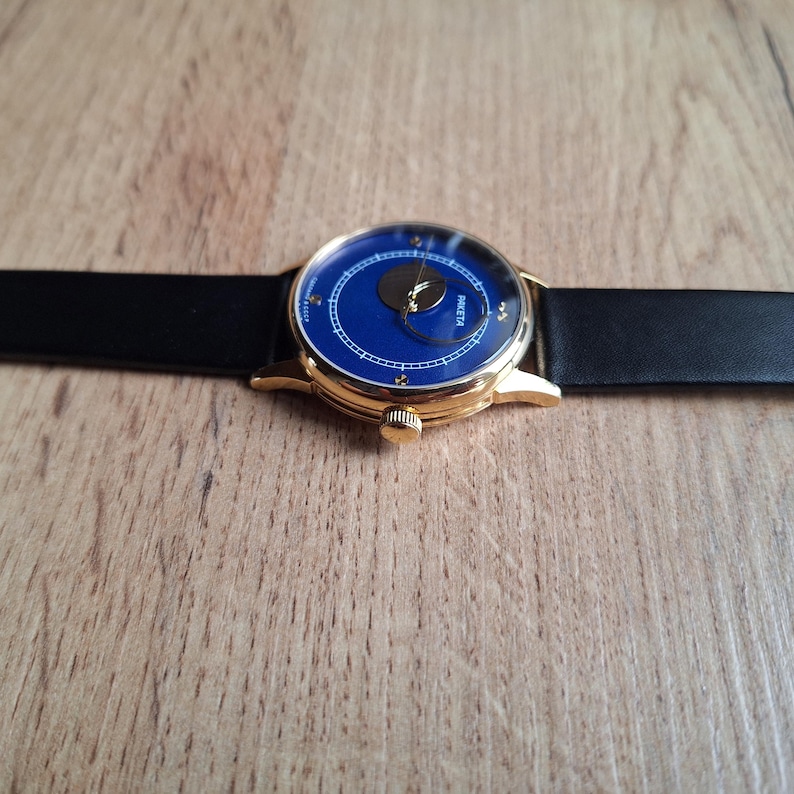 Op de afbeelding: Een gouden polshorloge met een blauwe wijzerplaat en een zwarte leren band. De wijzerplaat van het horloge heeft een maanfase-complicatie en het woord "PARETA" erop gedrukt.