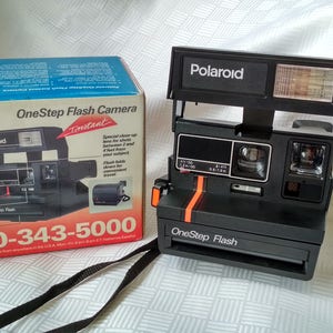 Peut inclure: Un appareil photo instantané Polaroid OneStep Flash noir avec une bande orange et un flash blanc. L'appareil photo est posé sur une surface blanche. La boîte de l'appareil photo est bleue et blanche et porte l'inscription "Polaroid OneStep Flash Camera Instant" sur le devant.