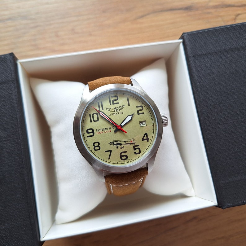 K&ouml;nnte beinhalten: Eine braune Lederarmbanduhr mit einem silbernen Geh&auml;use und einem wei&szlig;en Zifferblatt. Das Zifferblatt der Uhr hat schwarze Zahlen und Zeiger, einen roten Sekundenzeiger und ein kleines Datumsfenster bei 3 Uhr. Das Zifferblatt der Uhr zeigt auch eine kleine Flugzeuggrafik und den Text "Авиатор" (Aviator) und "Гастелло Н. Ф. Герой СССР" (Gastello N. F. Hero of the USSR).