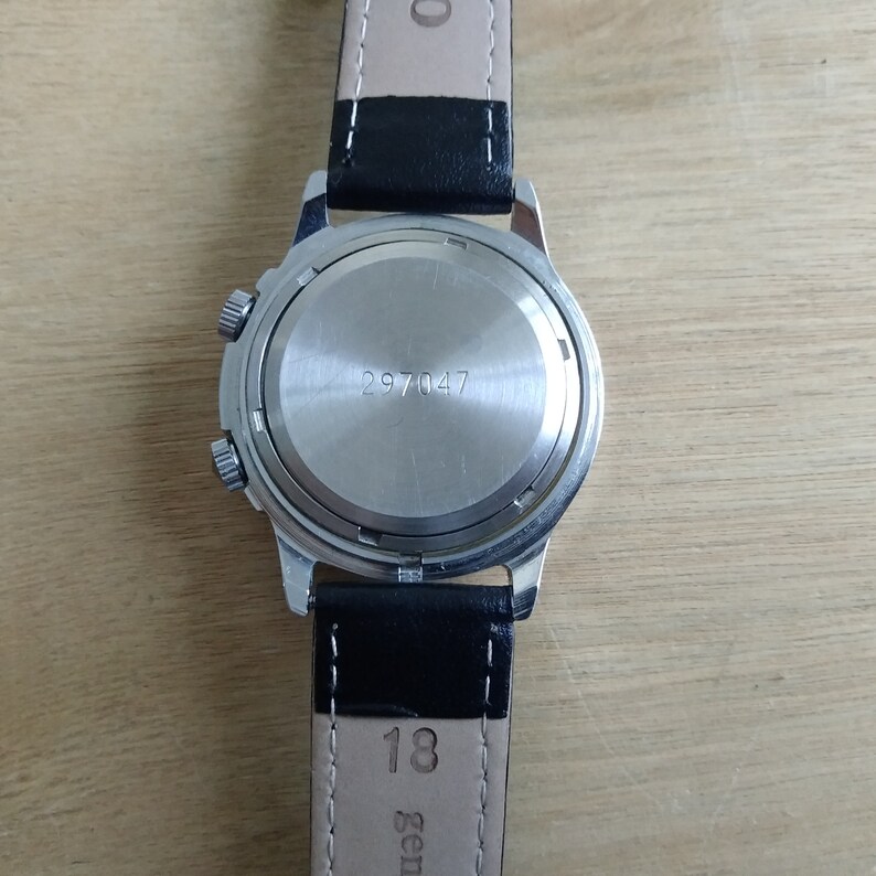 Puede incluir: La parte posterior de un reloj de pulsera plateado con una correa de cuero negro. La esfera del reloj tiene una caja plateada con el n&uacute;mero 297047 impreso en ella. La correa tiene el n&uacute;mero 18 impreso en ella.