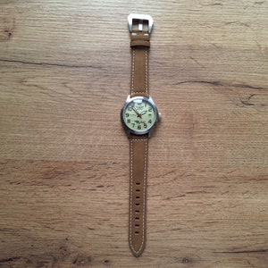 K&ouml;nnte beinhalten: Eine braune Lederarmbanduhr mit wei&szlig;em Zifferblatt und silbernen Zeigern. Die Uhr hat ein wei&szlig;es N&auml;hdetail am Rand des Armbands.