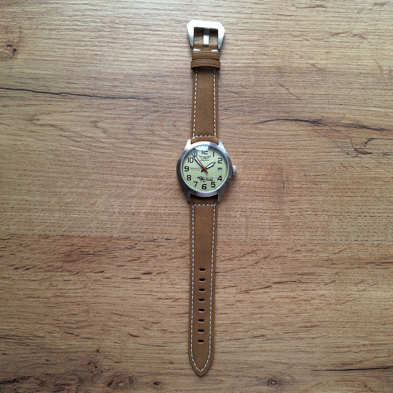 K&ouml;nnte beinhalten: Eine braune Lederarmbanduhr mit wei&szlig;em Zifferblatt und silbernen Zeigern. Die Uhr hat ein wei&szlig;es N&auml;hdetail am Rand des Armbands.
