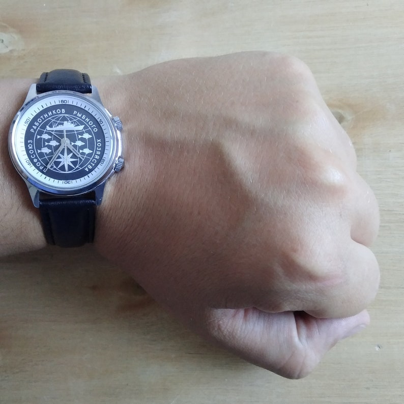 Puede incluir: Un reloj de pulsera plateado con una correa de cuero negro. La esfera del reloj tiene un fondo blanco con texto negro y un dise&ntilde;o de br&uacute;jula. El texto en la esfera del reloj dice "Работников Рыбного Хозяйства СССР".