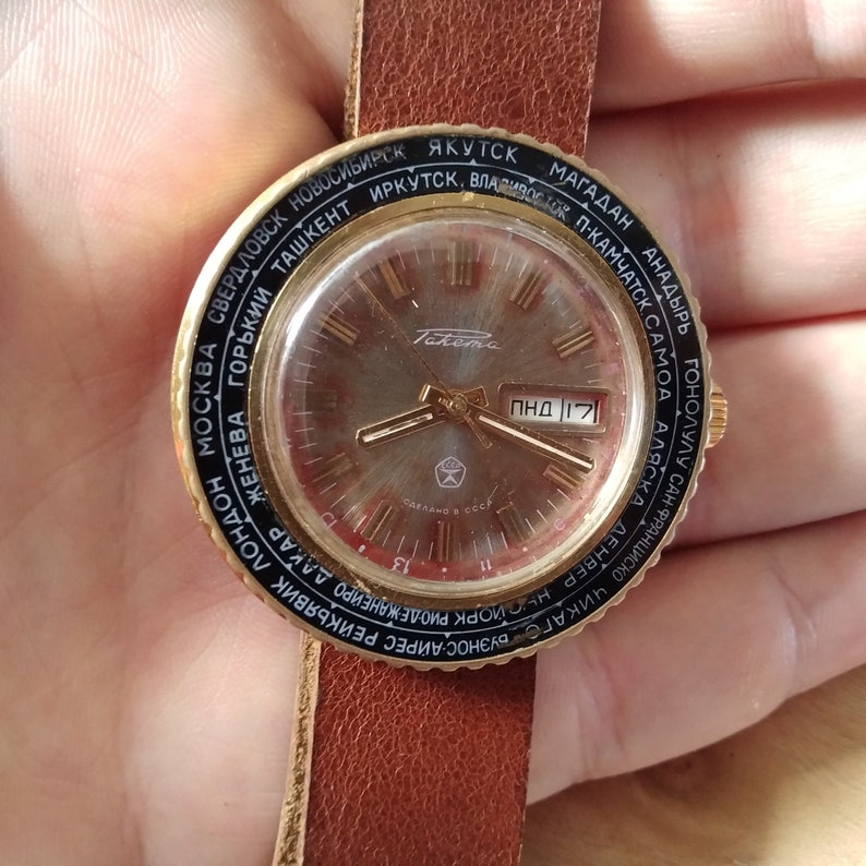Puede incluir: Un reloj de pulsera dorado con una correa de cuero negro. La esfera del reloj tiene un fondo plateado con marcadores de hora en n&uacute;meros romanos negros y una ventana de fecha que muestra "ПНД 17". La esfera del reloj est&aacute; rodeada por un anillo negro con nombres de ciudades impresos en blanco.