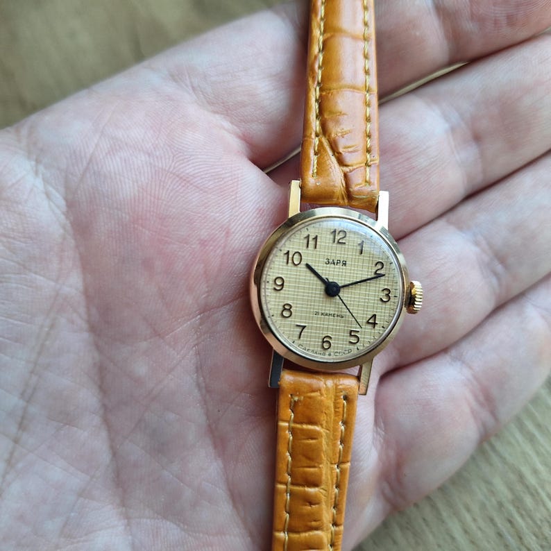 Op de afbeelding: Een vintage goudkleurig polshorloge met een lichtbruine leren band. De wijzerplaat toont zwarte cijfers en wijzers op een cr&egrave;mekleurige achtergrond. De band is ongeveer 1,3 cm breed.