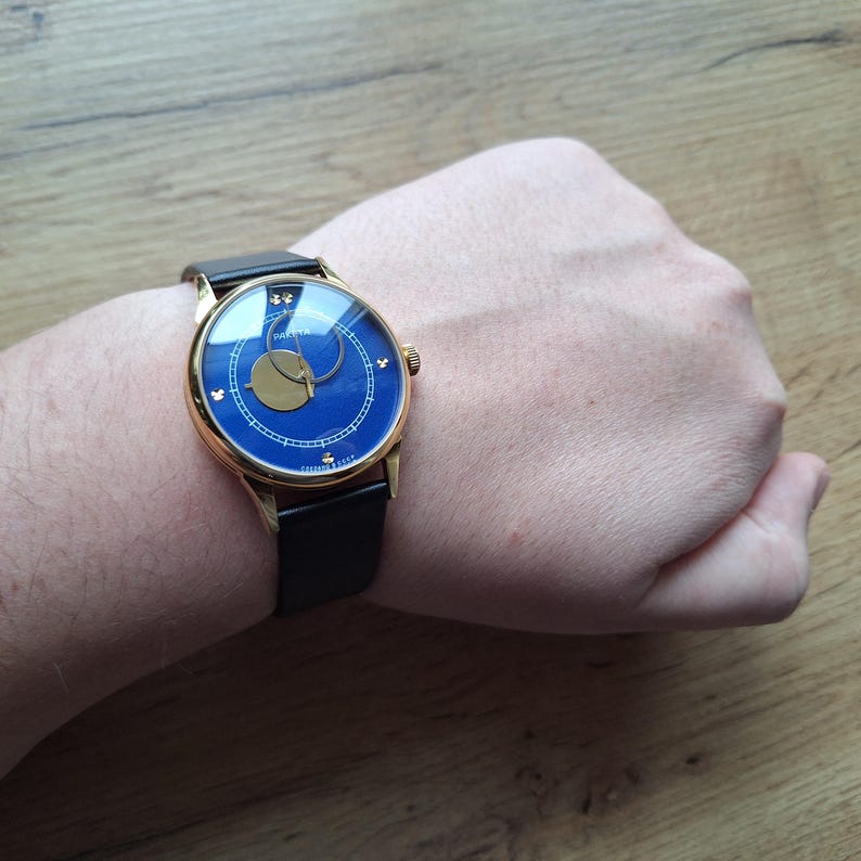 Op de afbeelding: Een gouden polshorloge met een blauwe wijzerplaat en een zwarte leren band. De wijzerplaat van het horloge heeft een gouden maanfase-indicator en gouden wijzers.