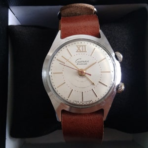 Op de afbeelding: Een zilveren polshorloge met een witte wijzerplaat en zwarte wijzers. Het horloge heeft een bruin leren bandje en staat "Слава 18 камней" op de wijzerplaat.