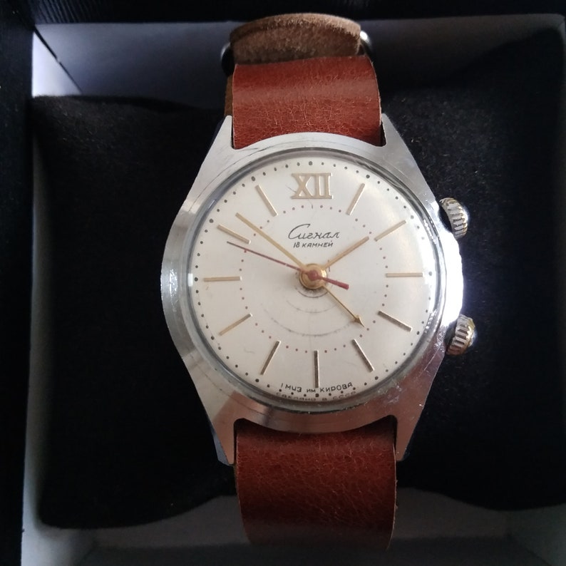 Op de afbeelding: Een zilveren polshorloge met een witte wijzerplaat en zwarte wijzers. Het horloge heeft een bruin leren bandje en staat "Слава 18 камней" op de wijzerplaat.