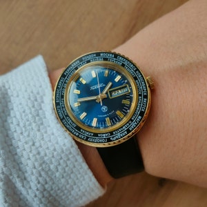 May include: A gold-tone wristwatch with a blue face and a black leather band. The watch face features a 24-hour time zone display with city names around the outer edge. The watch face also includes a date window and a day of the week window. The watch is made by the brand "Raketa".